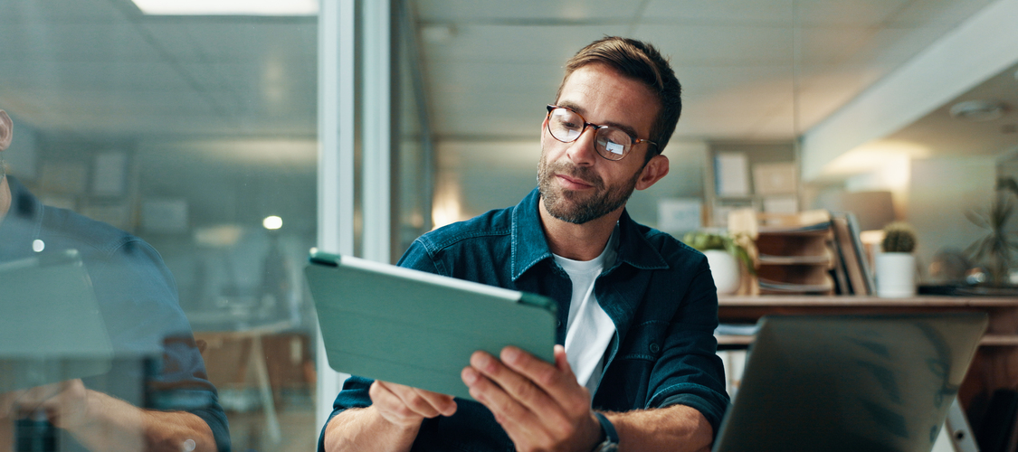 A man at a desk holding a tablet with a laptop open in front of him.