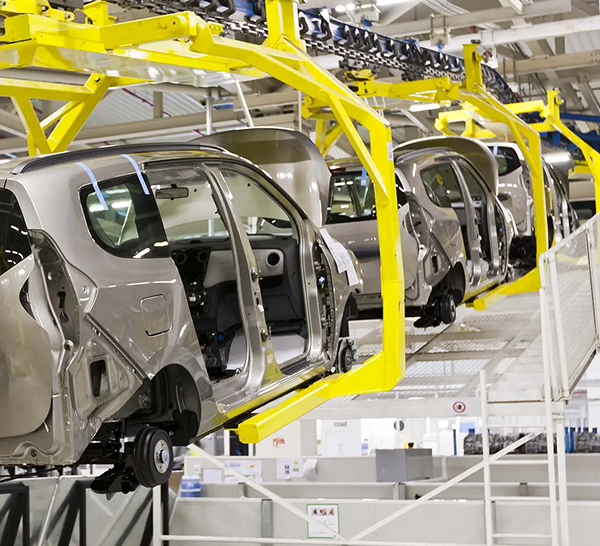 An image inside an automotive factory showing a line of unfinished car bodies, painted a metallic taupe color, moving along an overhead conveyor system. The car bodies are hanging from bright yellow metal framework. The scene illustrates an assembly line in a manufacturing environment.