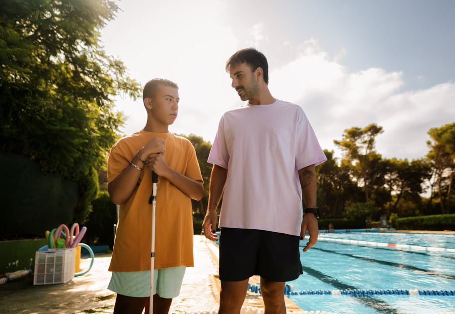 A blind young man and his friend walking and talking poolside on a sunny day.