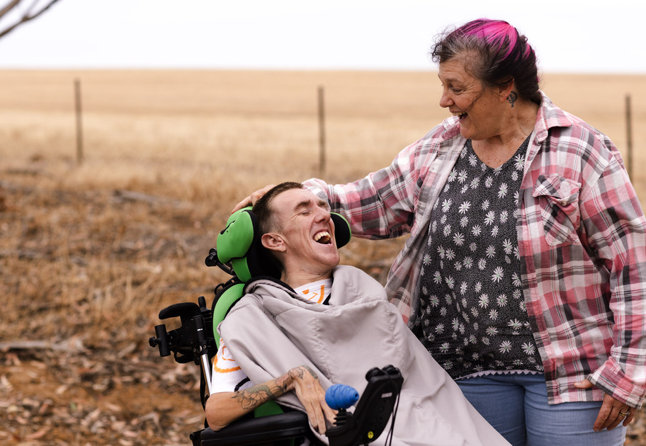 A son with quadriplegia and his mother outside by the road, talking and smiling together.