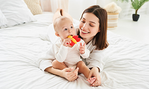 Mama na postelji z dojenčkom, ki izraščanje mlečnih zob lajša z grizalom