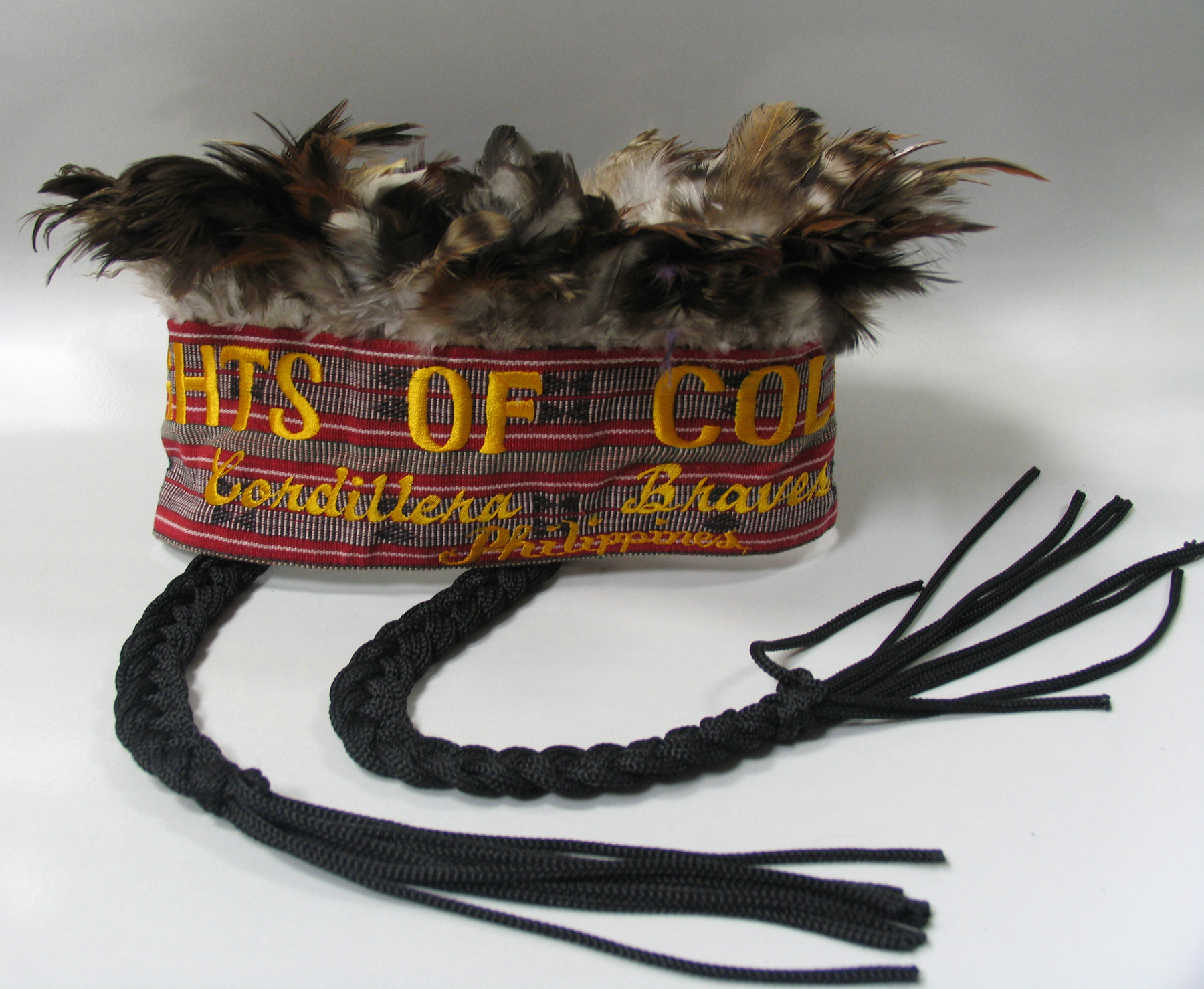 Contemporary headdress crowned with feathers and two hanging braids. Headband embroidered with text “Knights of Columbus Cordillera Braves Phillippines.”