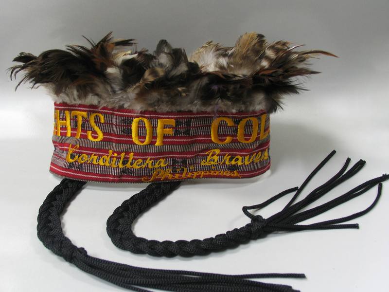 Contemporary headdress crowned with feathers and two hanging braids. Headband embroidered with text “Knights of Columbus Cordillera Braves Phillippines.”