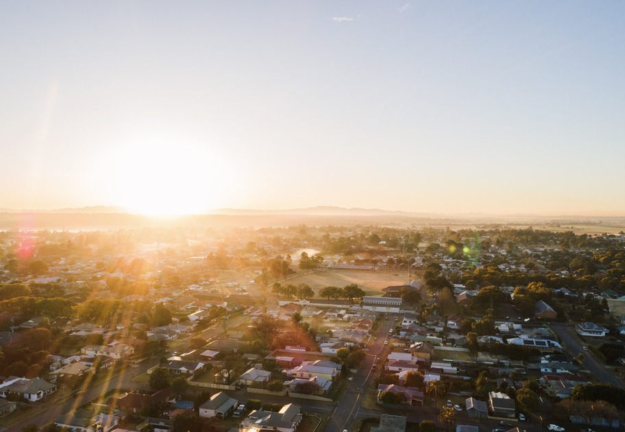Aerial view of a suburban neighborhood at sunrise, with sunlight casting a warm glow over houses, trees, and distant hills under a clear sky.