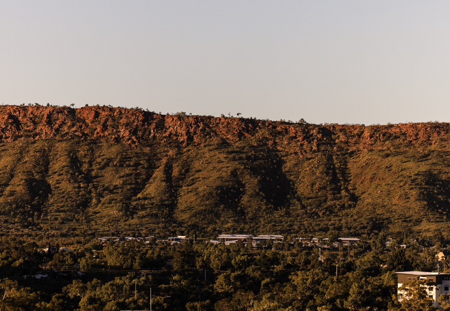 A rocky, sunlit ridge rises above a small town nestled among dense green trees, under a clear sky.