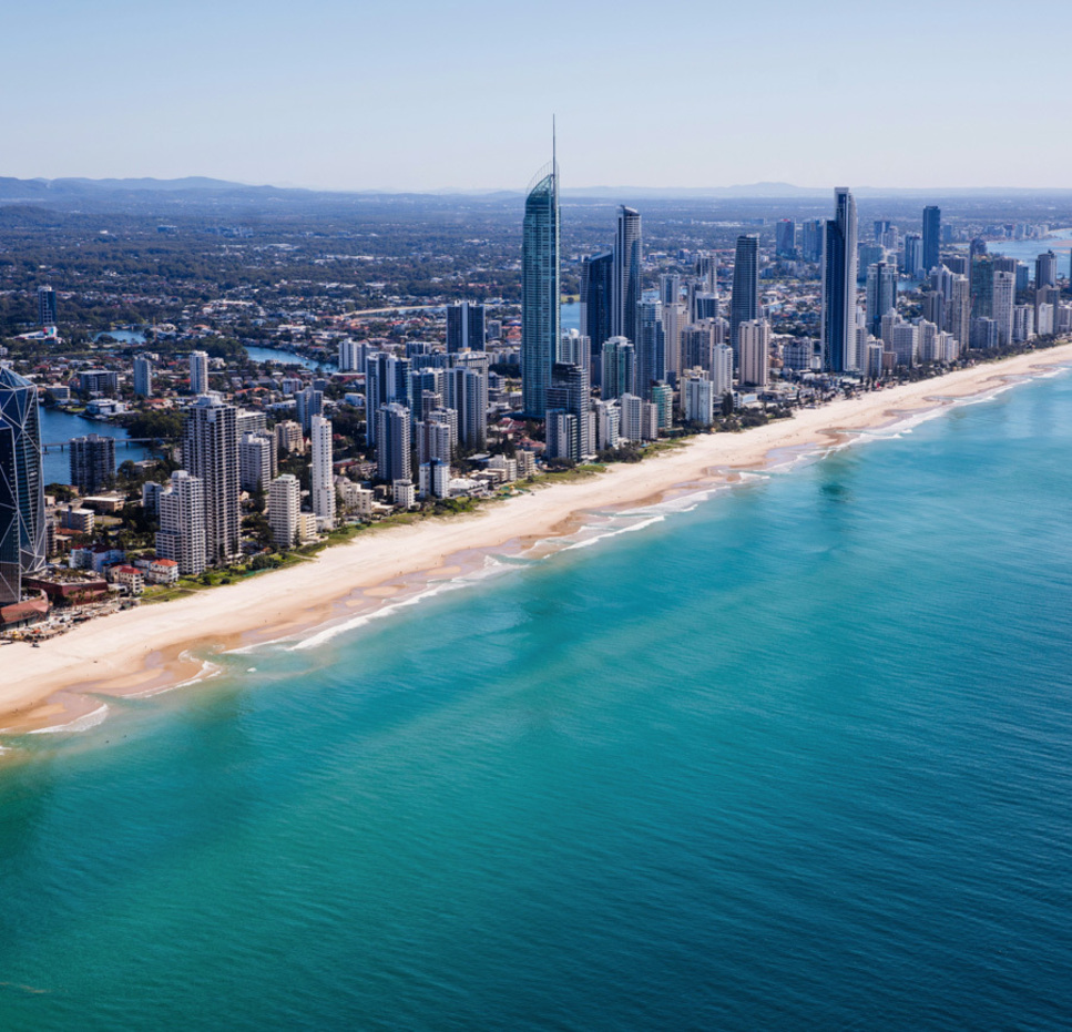 Aerial view of Gold Coast skyline with tall buildings lining a long sandy beach, clear turquoise ocean water, and mountains in the distant background under a clear blue sky.