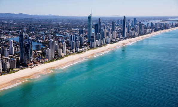 Aerial view of Gold Coast skyline with tall buildings lining a long sandy beach, clear turquoise ocean water, and mountains in the distant background under a clear blue sky.