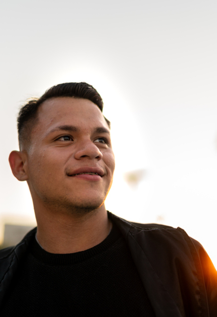 A young man with short dark hair smiles slightly while looking to the side. He is outdoors during sunset, wearing a black jacket and shirt, with blurred trees and palm trees in the background.
