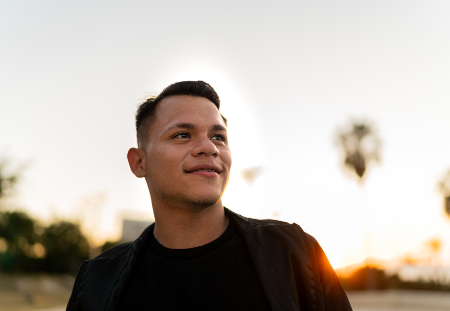 A young man with short dark hair smiles slightly while looking to the side. He is outdoors during sunset, wearing a black jacket and shirt, with blurred trees and palm trees in the background.