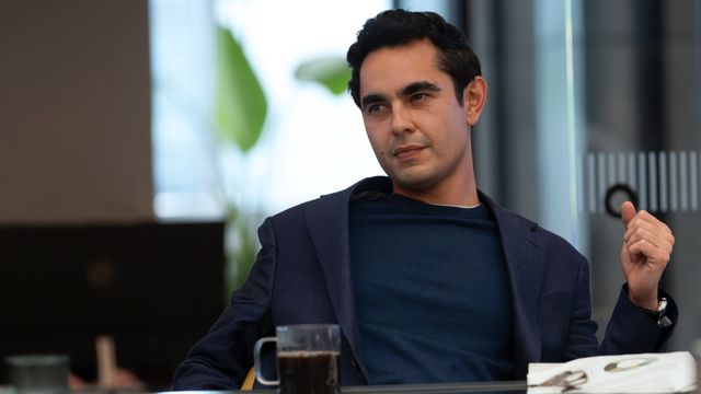 Max Minghella as Whitney Halberstram sitting at a desk with coffee, gesturing while conversing to someone out of frame