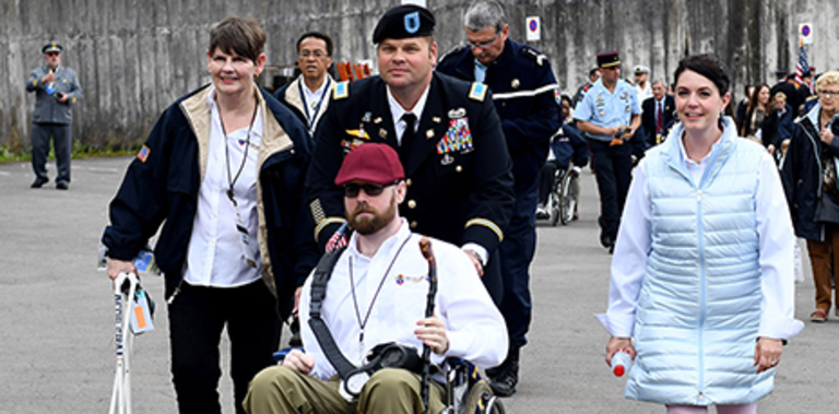 Charles sits in a wheelchair in the center, surrounded by his wife and military pilgrims. A man in military uniform pushes the wheelchair