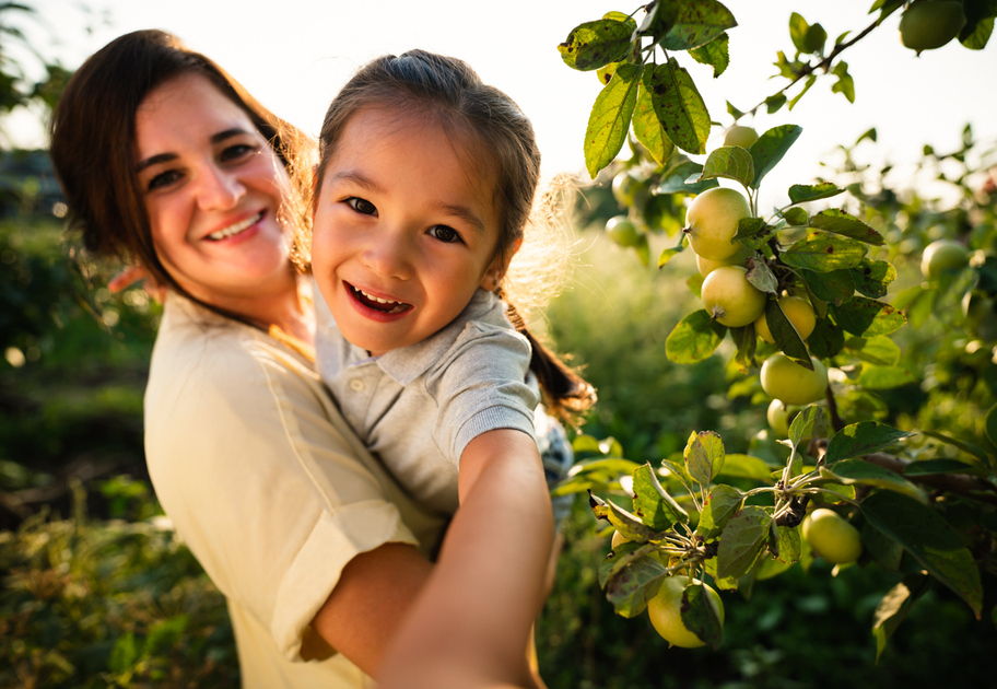 A smiling woman holds a cheerful young girl in an orchard, with green apples growing on a tree nearby. Sunlight shines behind them, highlighting their happy expressions.