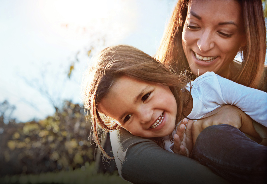 A mother hugs her daughter outside in the summer sunshine. They are sharing a warm embrace. 