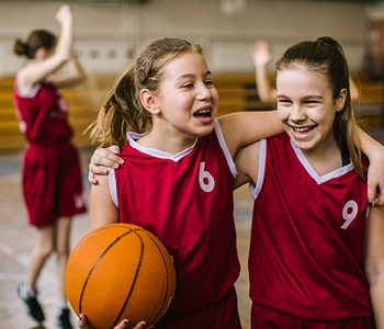 Dve deklici v rdečih košarkarskih uniformah stojita objeti v telovadnici pri obšolski dejavnosti.