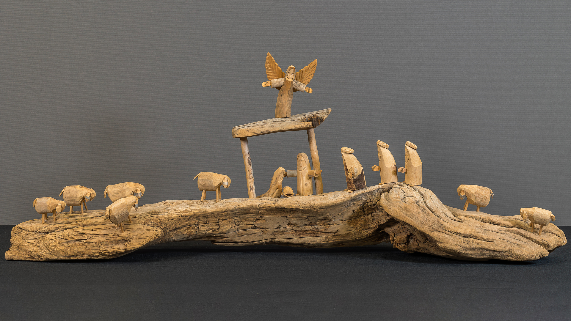 Photo of a crèche featuring the wood carved figures of the Holy Family in the stable, an Angel, the 3 Magi, and 7 sheep on a piece of driftwood. 