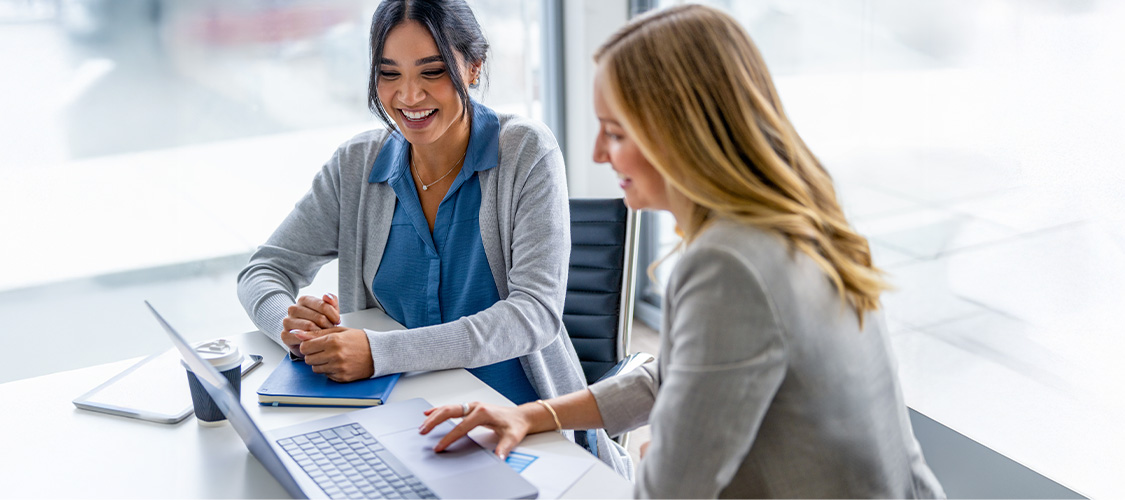 Two women sitting down smiling and talking and looking at a laptop.