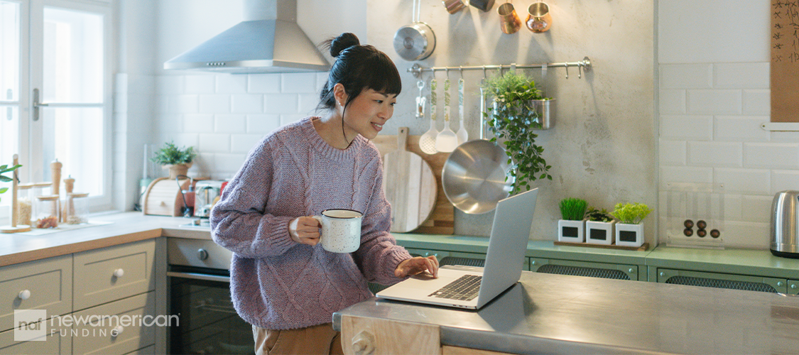 A woman in a kitchen looking at a laptop while holding a mug.