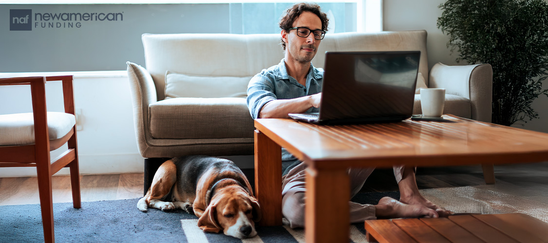 A man sitting on the floor typing on a laptop with a dog lying next to him.