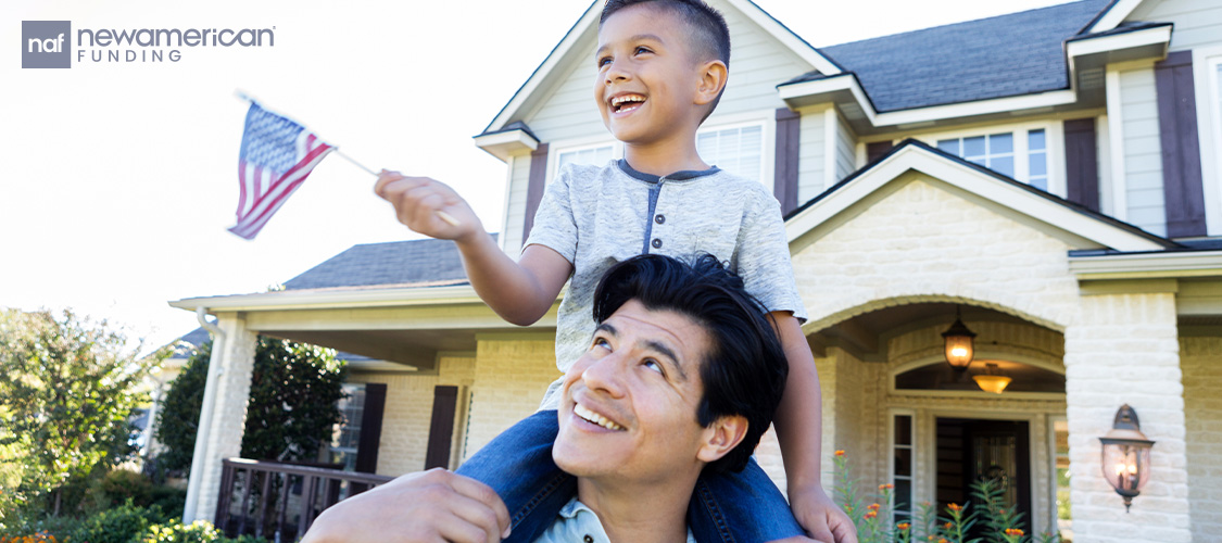 A small boy with a small American flag on the shoulders of a man in front of a house.