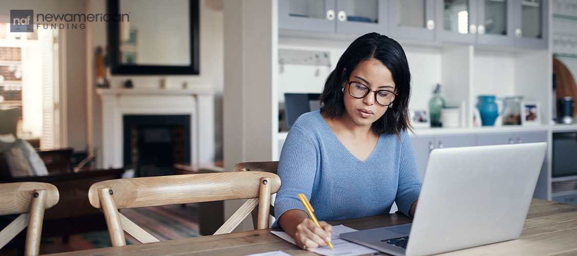 A woman sitting in front of a laptop and writes on a piece of paper next to her.