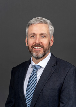 Headshot of Richard Shea, who smiles while wearing a black suit and a blue tie