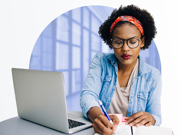 A focused woman wearing glasses and a denim shirt with a bandana is writing in a notebook while looking at a laptop screen, framed by a circular white cutout against a blurred blue office background.