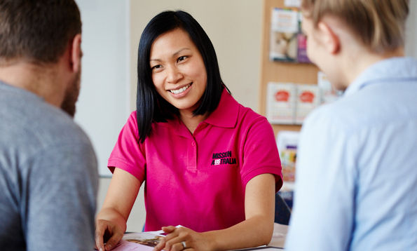 A Mission Australia case worker sits at a desk, smiling and talking with two people. She holds a brochure, and shelves with pamphlets are visible in the background.