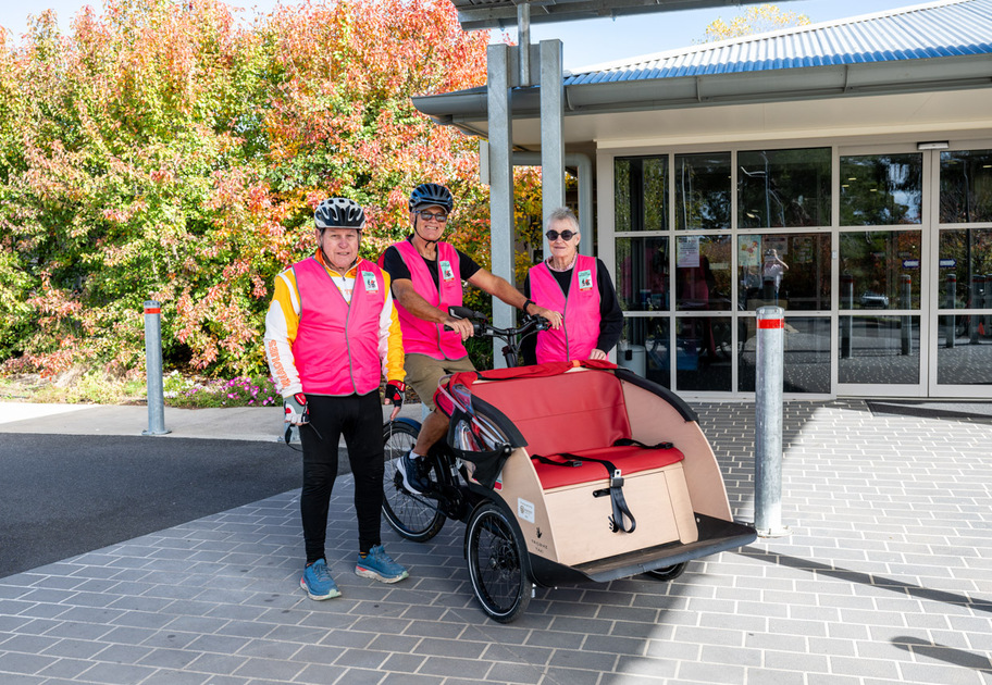 Three people wearing bright pink vests and bike helmets stand beside a special bicycle with a large front passenger seat outsideBenjamin Short Grove, with colorful autumn trees in the background.