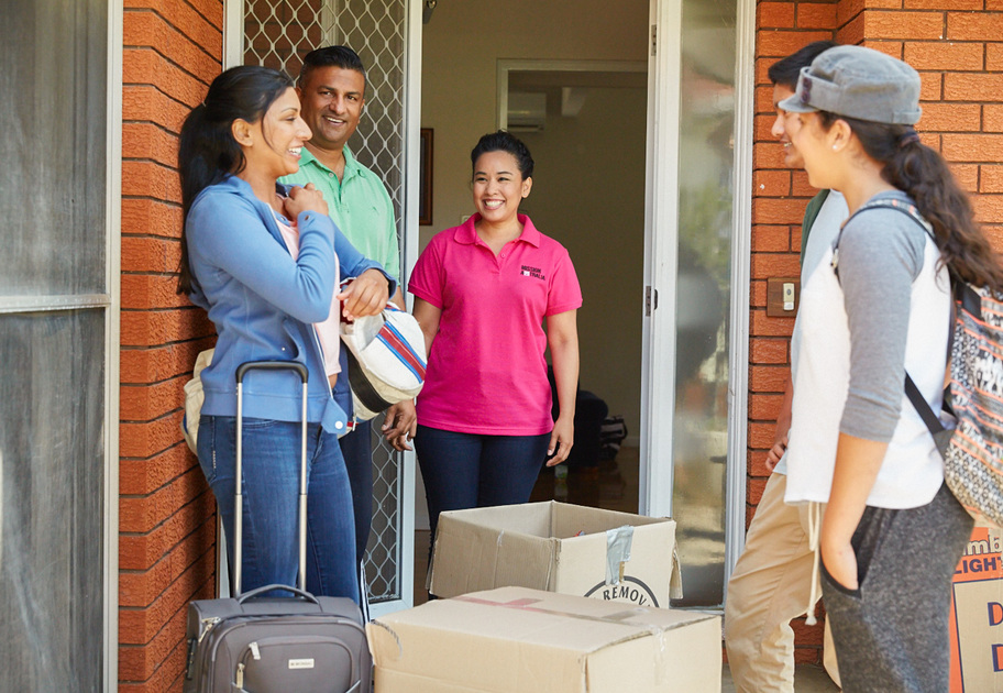 Three people stand at the door of a house, smiling and greeting a fourth person who is arriving with a backpack and suitcase. Cardboard boxes are stacked by the doorway. It appears to be a welcoming or moving-in scene.