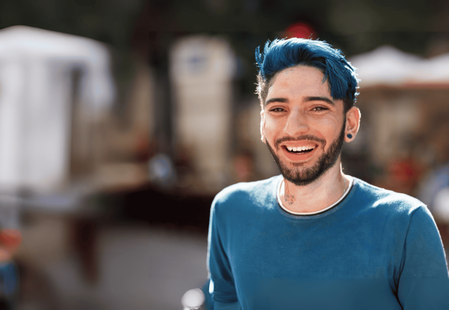 Smiling young person with bright blue hair and beard, wearing a teal shirt, standing outdoors in a sunny setting.