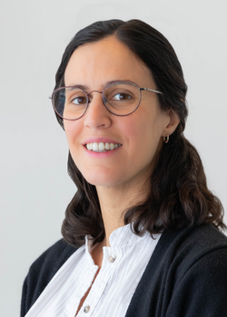 A corporate headshot style portrait of Sofia Maurette, smiling wearing a white blouse and black sweater on a light grey background.