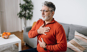 Starejši moški v rdečem puloverju sedi na kavču in zdi se, da občuti znake srčnega infarkta – bolečino v prsih in slabost.