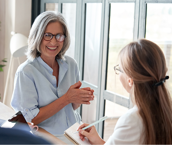 A mature woman with grey hair and glasses, wearing a light blue shirt, smiles and gestures with her hands while talking to a person taking notes across a table in a bright room.
