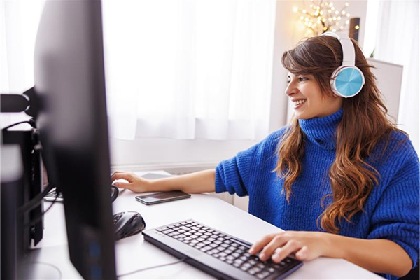 A young woman with long, brown hair and a bright blue turtleneck sweater smiles while sitting at a desk and working on a computer. She is wearing light blue and white over-ear headphones. She is looking at the screen, and her hands are on the keyboard and mouse.