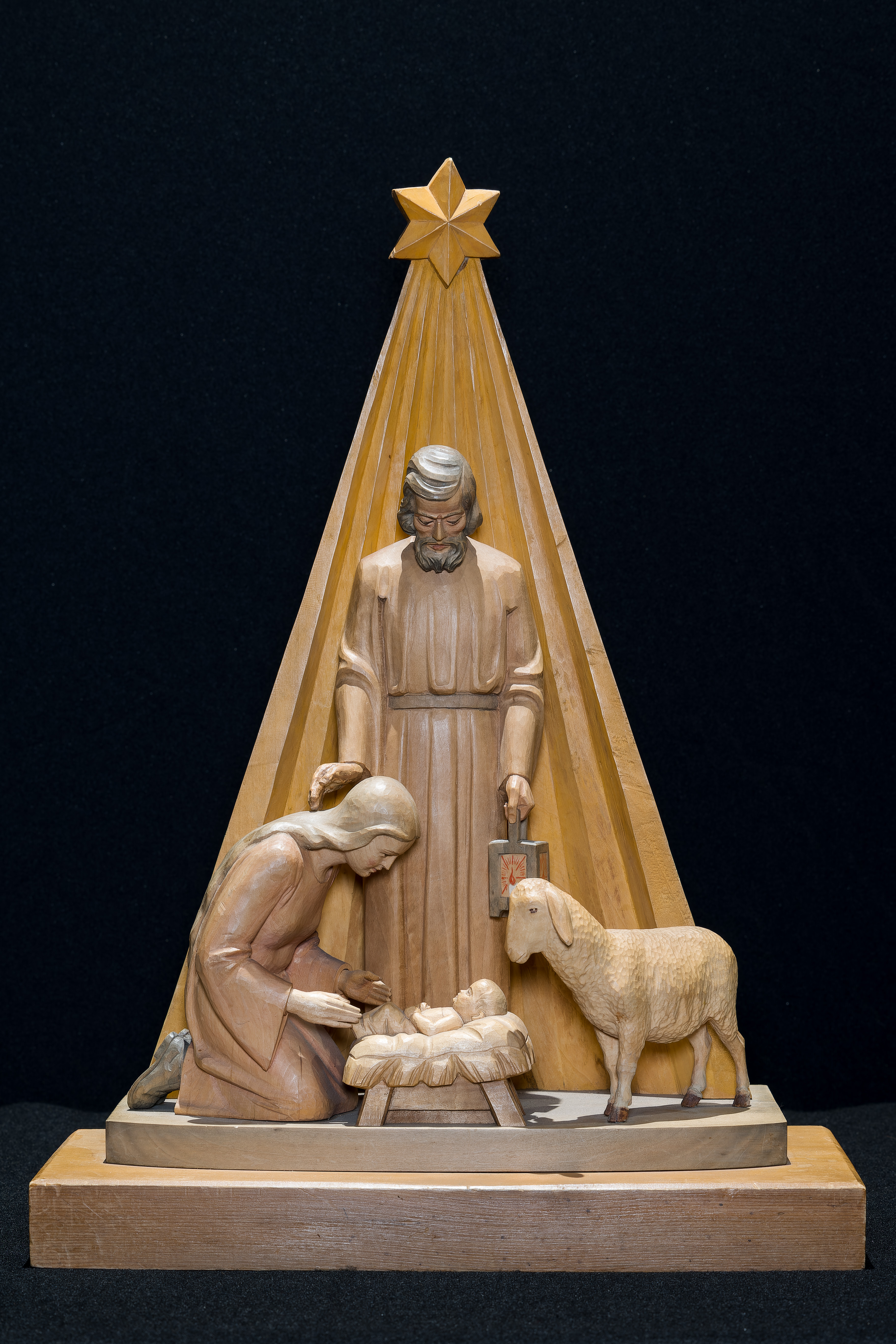 Image of a wood carved Nativity featuring the Holy Family and a sheep standing before a star and its radiating beams. 