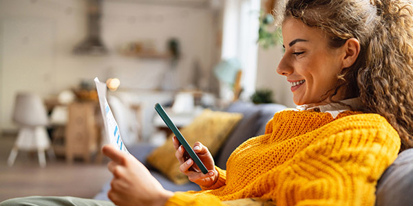 A smiling woman looks at her phone screen and a piece of paper in her hand