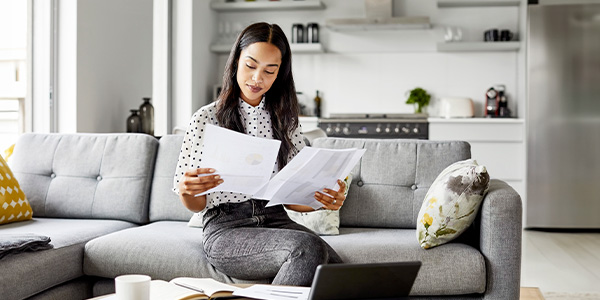 A Black woman sits on a couch in a home holding and comparing two sheets of paper with information on them