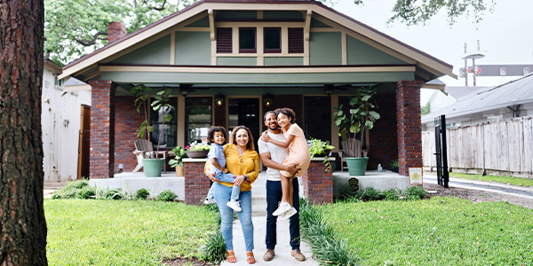 A smiling Latina woman stands with a smiling Black man and two smiling children in front of a nice home