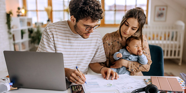 A white man and woman sit at a table looking at financial forms while the woman holds a baby