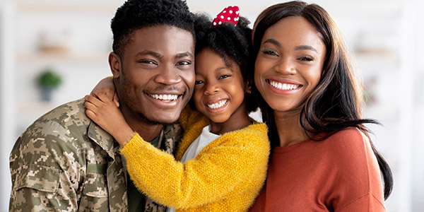 A Black man in a military uniform smiles into the camera next to a smiling Black woman and child 