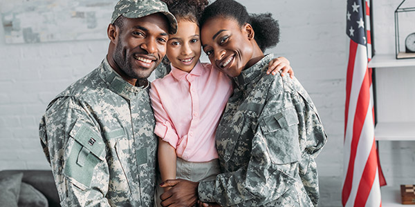 A Black man and Black woman dressed in military uniforms stand smiling on either side of a Black girl with an American flag behind them