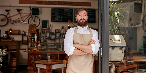 A white man stands in front of a shop wearing a white button down and a brown apron