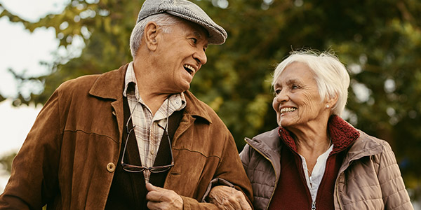A senior white man and woman smile at each other as they walk together