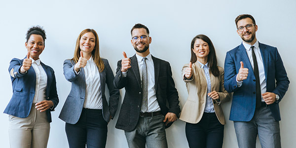 A diverse group of five people dressed in suits smile into the camera while giving a thumbs up