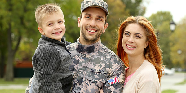 A white man stands smiling in a military uniform holding a young child with a smiling woman next to him