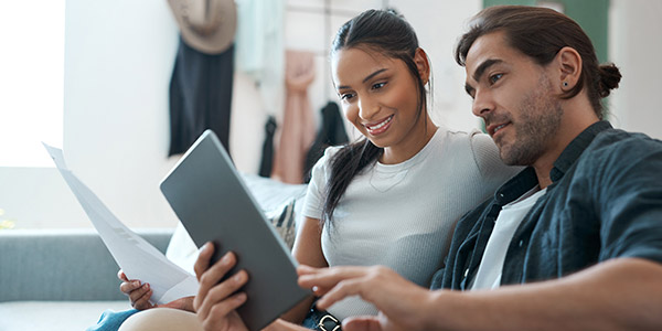 A Hispanic woman and man sit on the couch together looking at a tablet 
