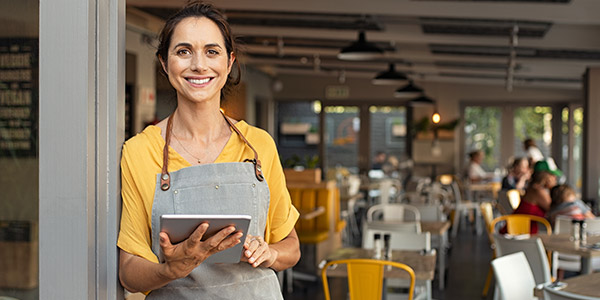 A Hispanic woman stands in front of a busy restaurant wearing an apron