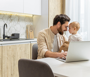 Mlad očka dela za računalnikom v domači kuhinji, z njim pa je 6-mesečni dojenček - prizor je ujel topel trenutek med njima.