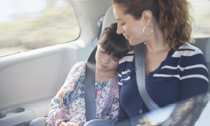 Mati in hči se vozita na zadnji klopi v avtomobilu. Hčerka spi z glavo na mamini rami, saj obe varuje varnostni pas.