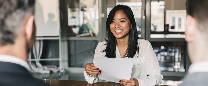 A photo of a woman with long dark hair, wearing a white shirt, seated at a table and holding papers, smiling while talking to two people seated opposite her, who are seen from the back. This setting suggests a meeting, interview, or registration process.